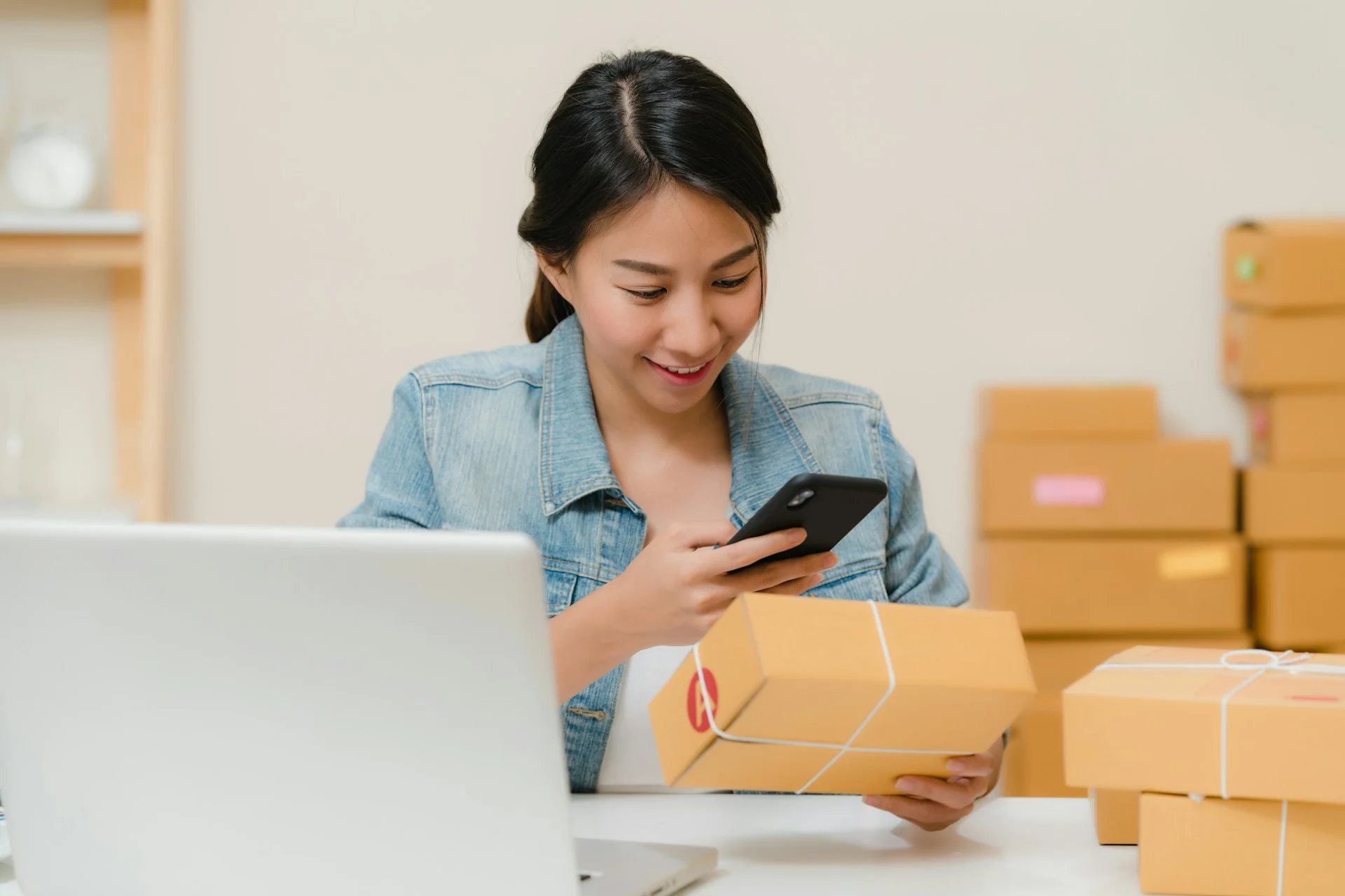Pemeriksaan paket pesanan — Google Shopping AI Seorang perempuan memeriksa paket dengan ponsel di dekat laptop dan kardus